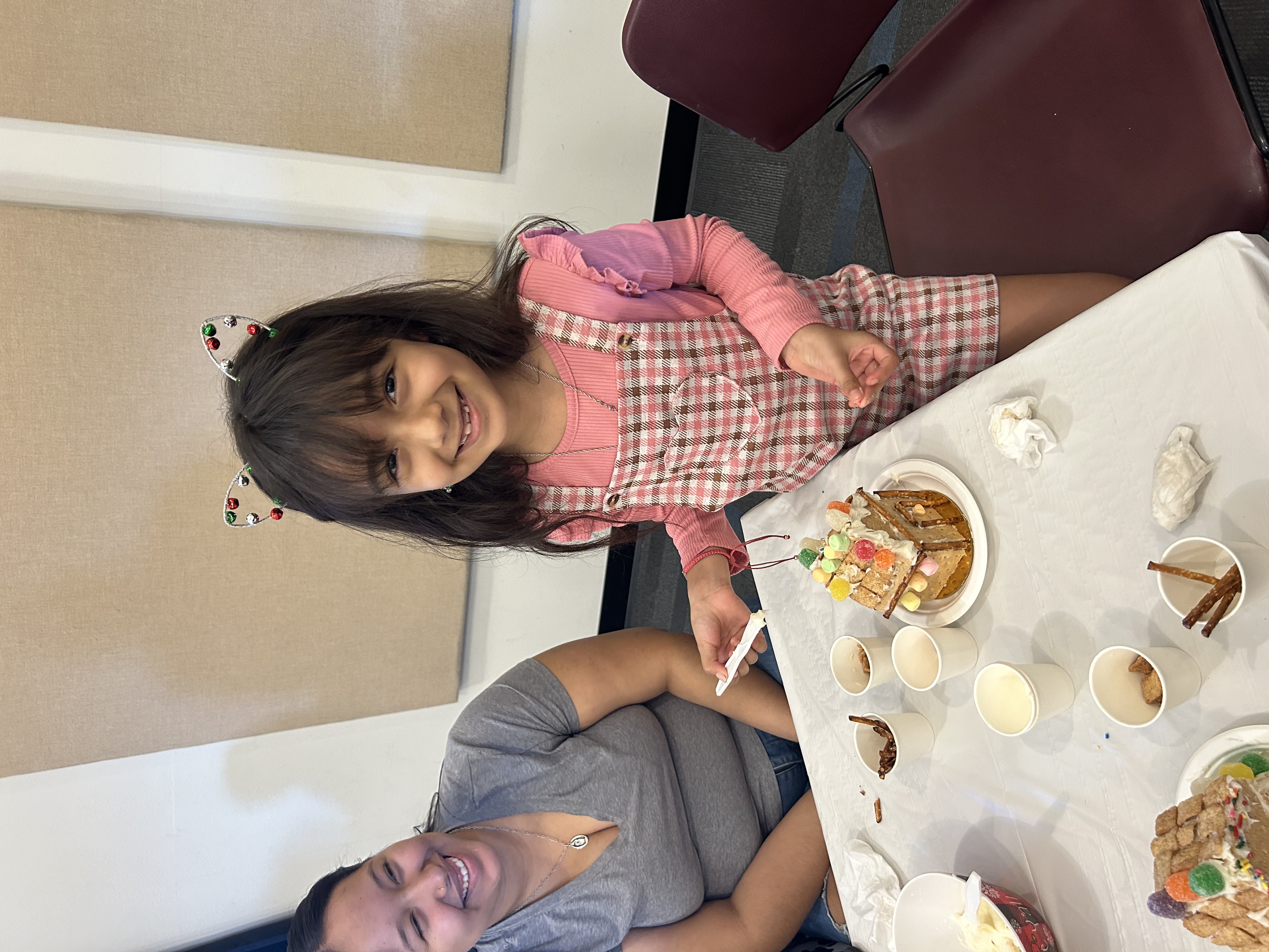 A smiling young girl wears cat ear headbands while decorating a gingerbread house with colorful candies. A woman beside her looks on cheerfully. The scene is joyful and festive.