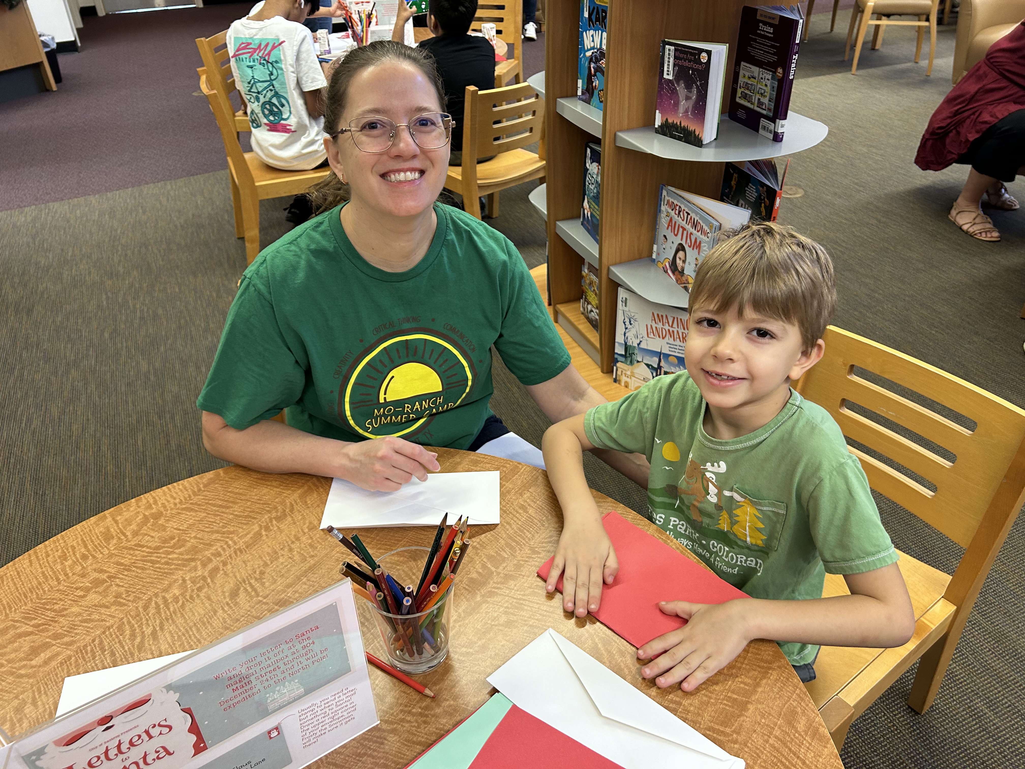 A smiling woman and a young boy sit at a round wooden table in a library, crafting with paper. Bookshelves and other patrons can be seen in the background.