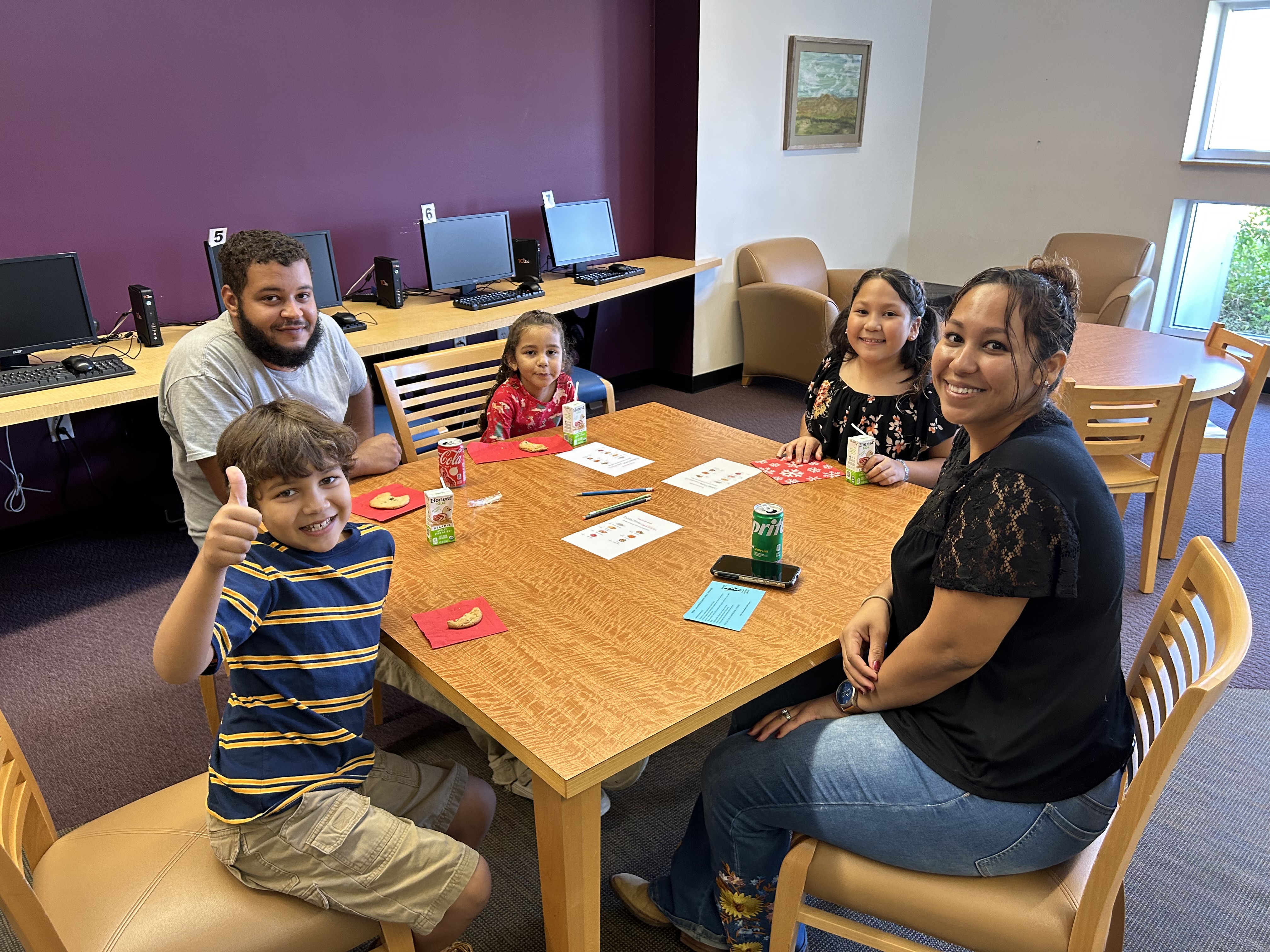 A group of people, including two adults and three children, sit happily around a wooden table in a library. One child gives a thumbs-up. Snacks and drinks are on the table, with computers in the background creating a warm, educational setting.