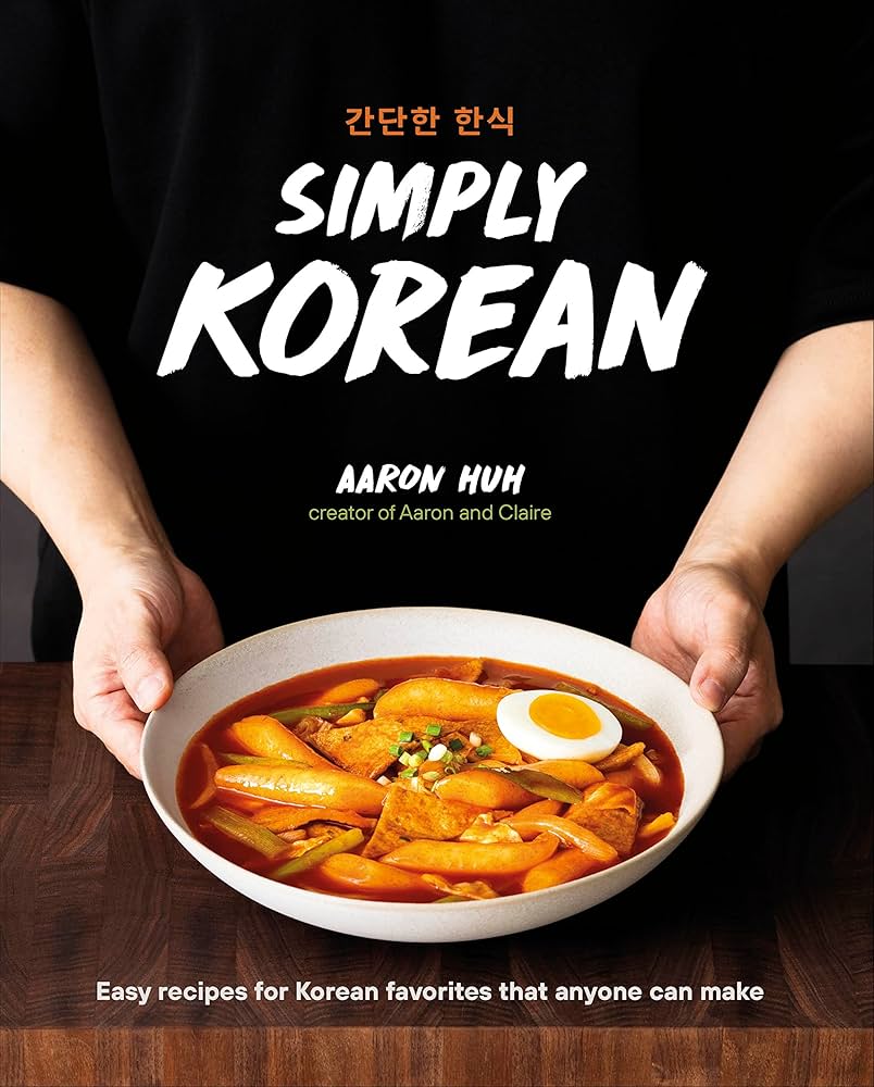 Hands holding a bowl of vibrant tteokbokki, a Korean dish with rice cakes, fish cakes, and boiled egg in spicy sauce. Text: "Simply Korean" by Aaron Huh.