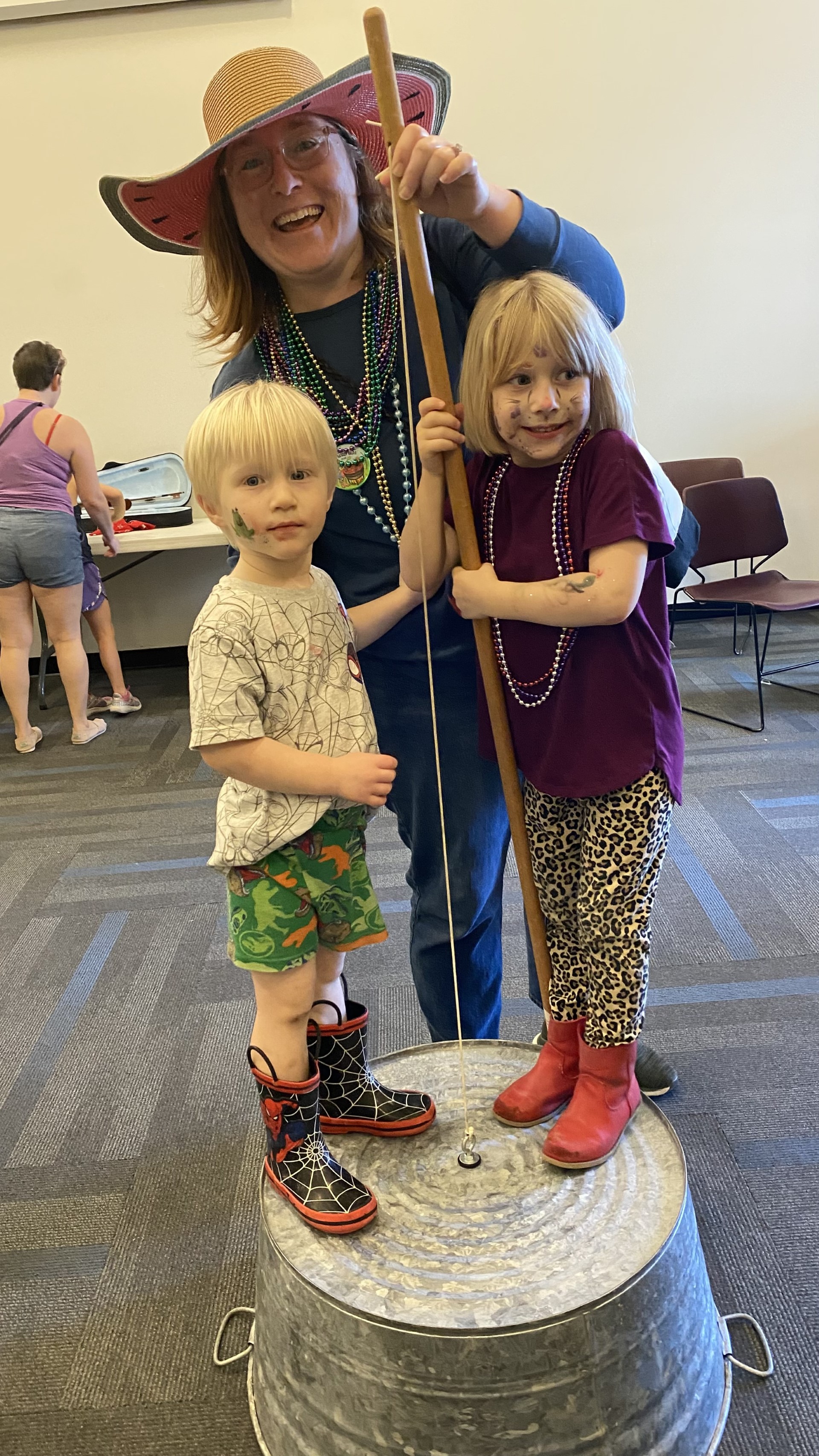A woman in a large floppy hat and beaded necklaces smiles behind two children standing on an upside-down metal tub, holding a wooden stick. Playful vibe.
