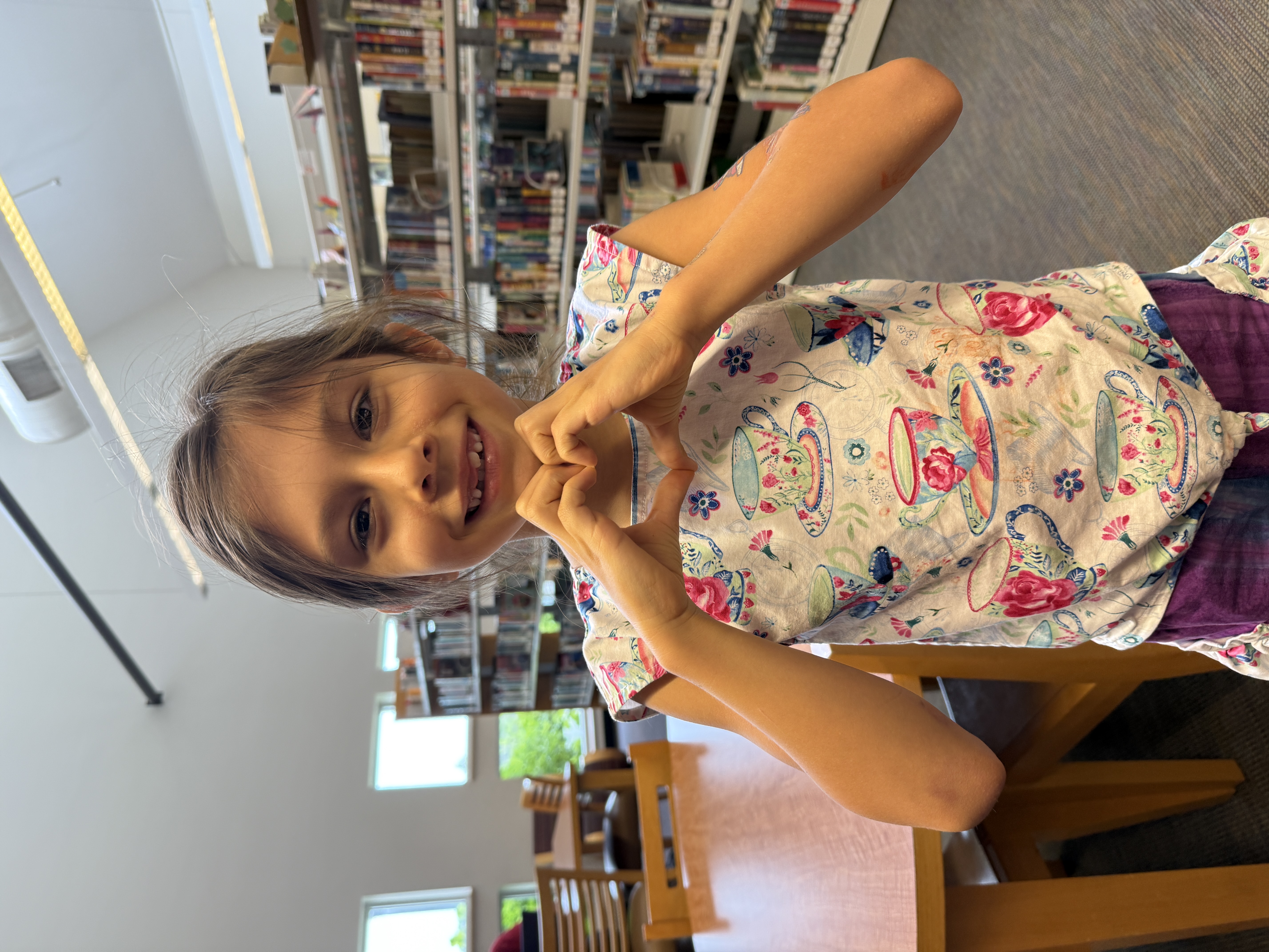 Smiling child in a teacup-patterned shirt forms a heart with hands, standing in a bright library. Bookshelves and tables are visible in the background.