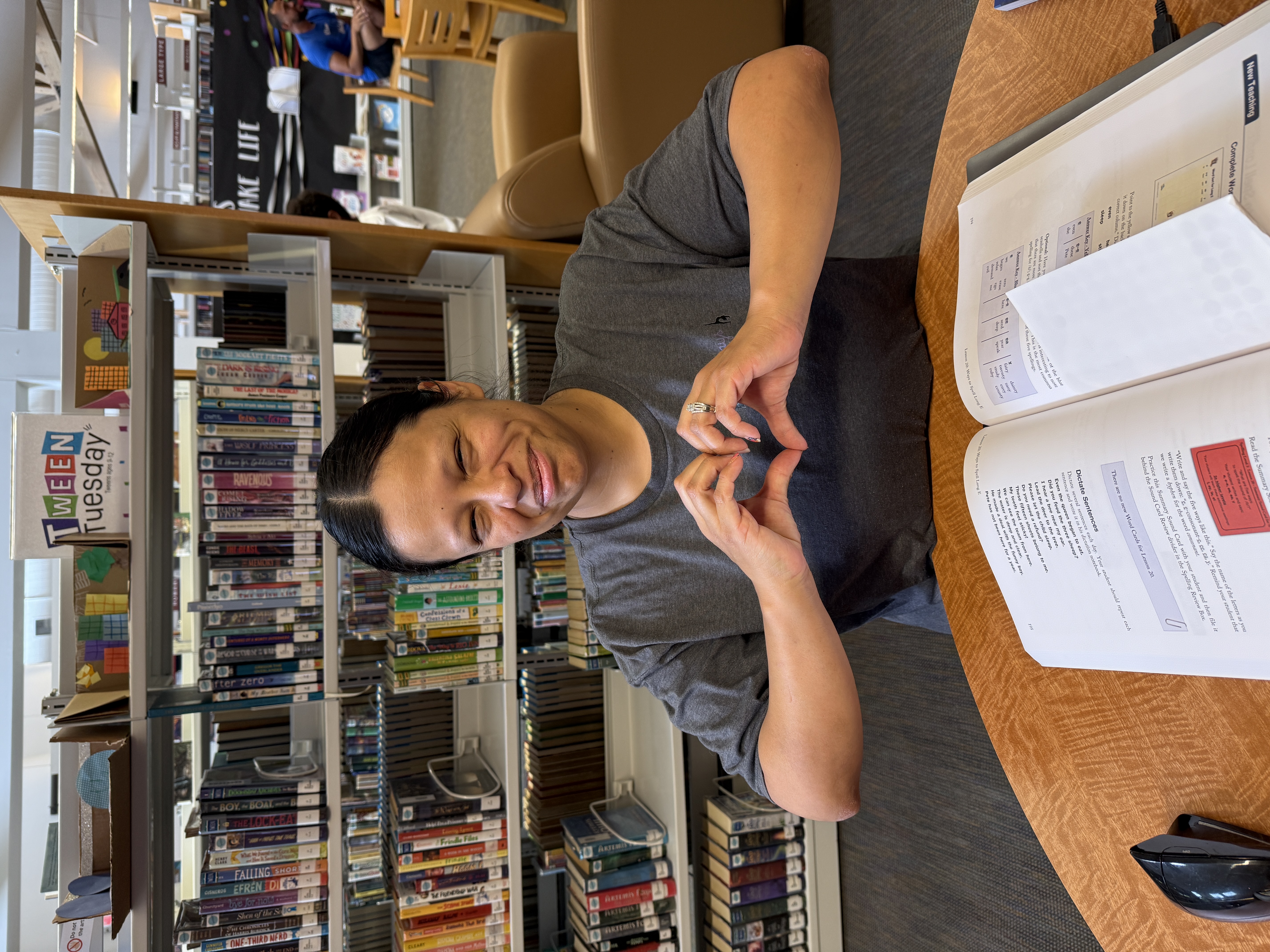 A person smiling while making a heart shape with their hands in a library. Open book on a table. "Tween Tuesday" sign on a bookshelf behind them.