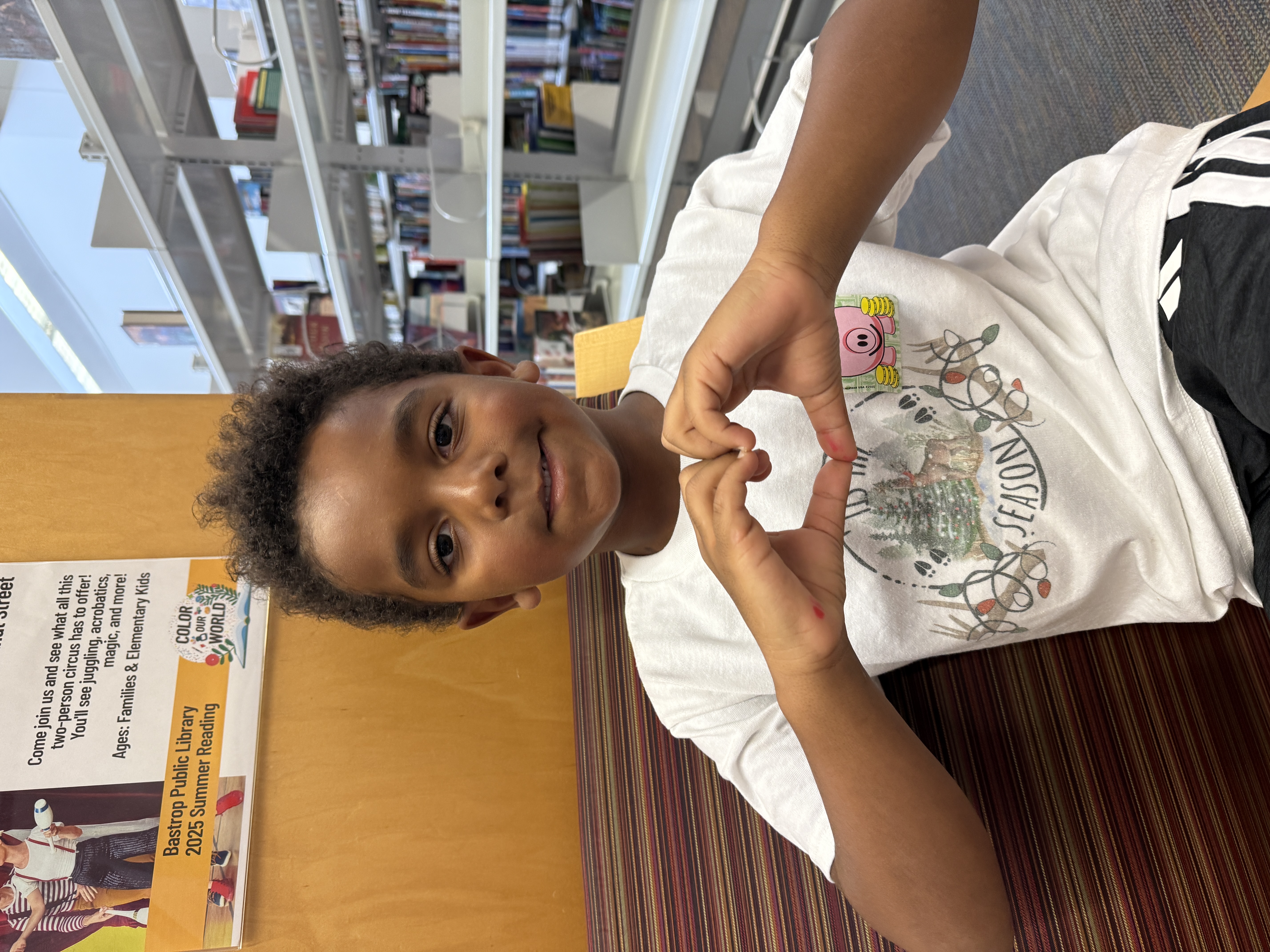 A young child in a library forms a heart shape with their hands, smiling warmly. The background features bookshelves and a colorful summer reading poster.