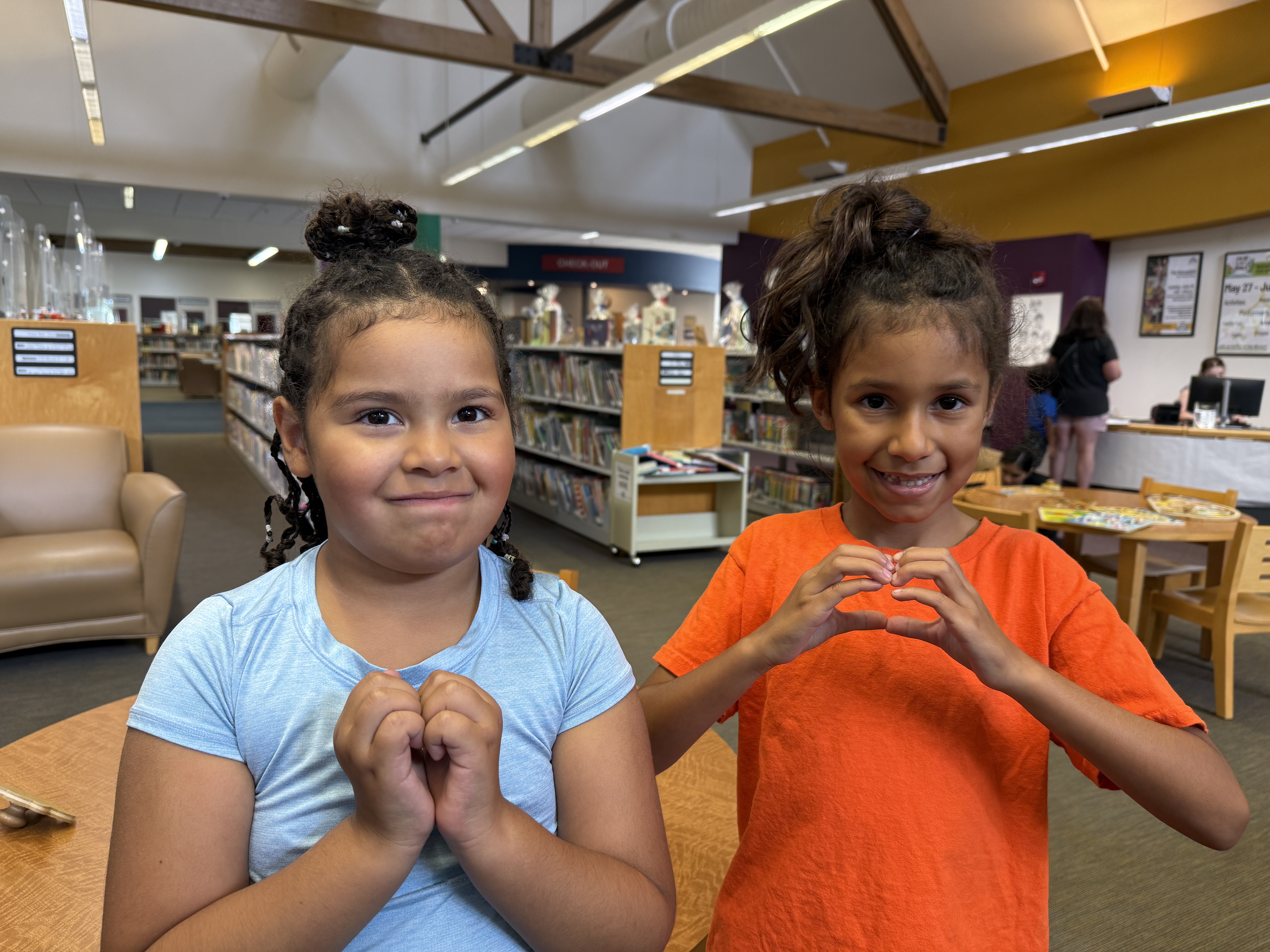 Two smiling young girls with dark hair stand in a bright library, forming heart shapes with their hands. One wears a blue shirt, the other orange.