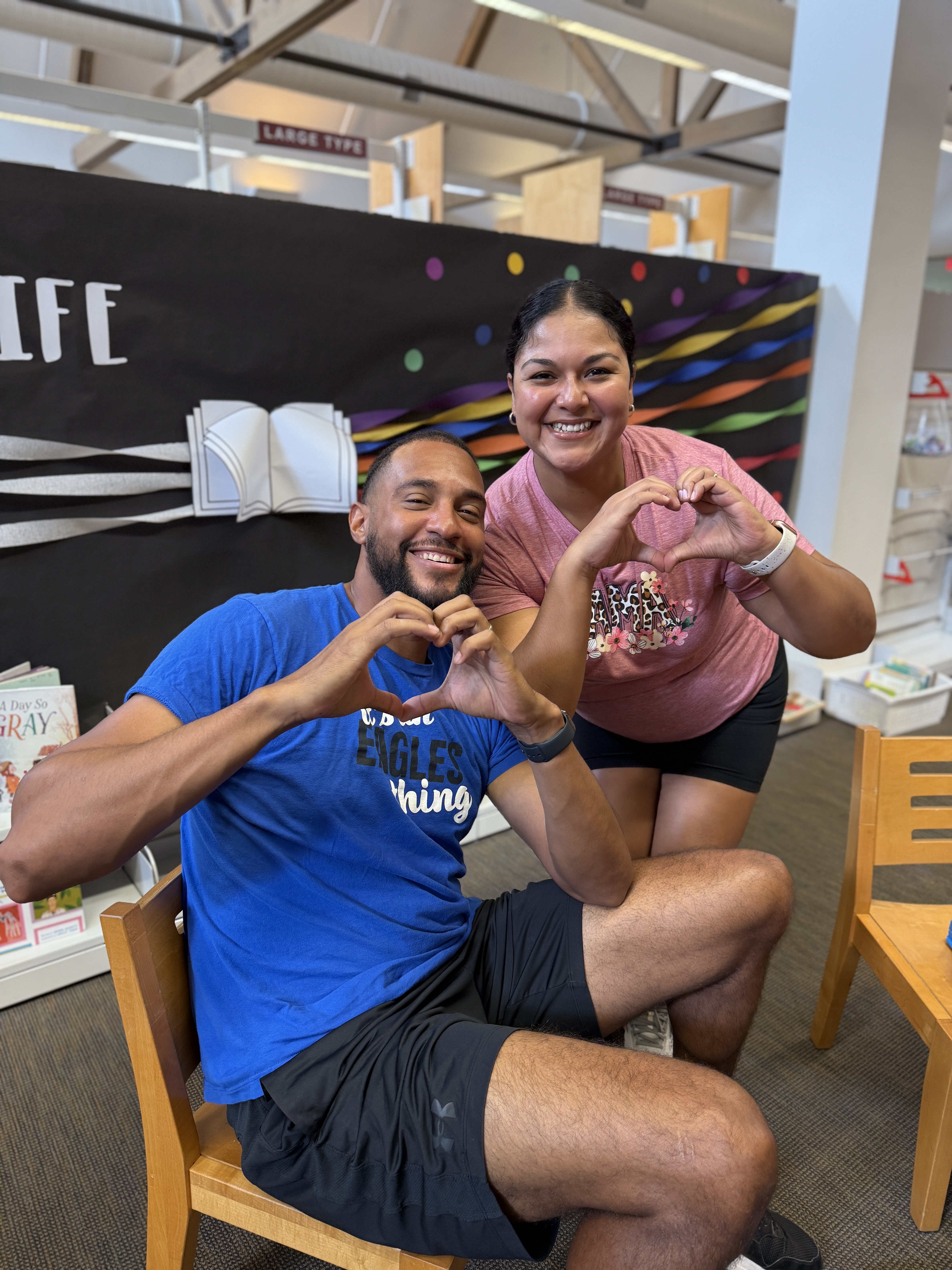 A smiling man and woman sitting on chairs in a library form heart shapes with their hands. The background features a blackboard with colorful stripes.