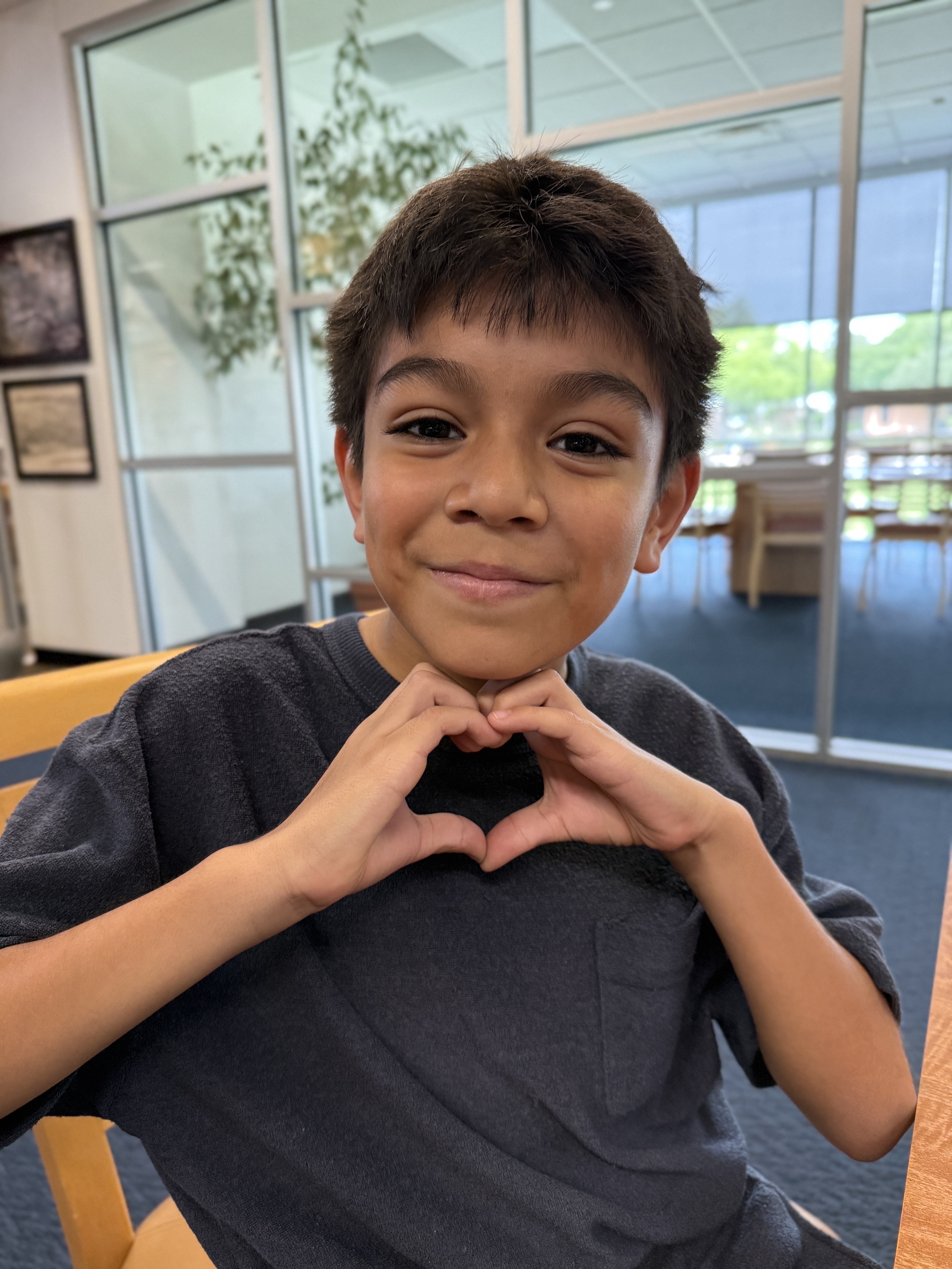 Young boy smiling and making a heart shape with his hands in a bright room with glass partitions and greenery, conveying warmth and friendliness.